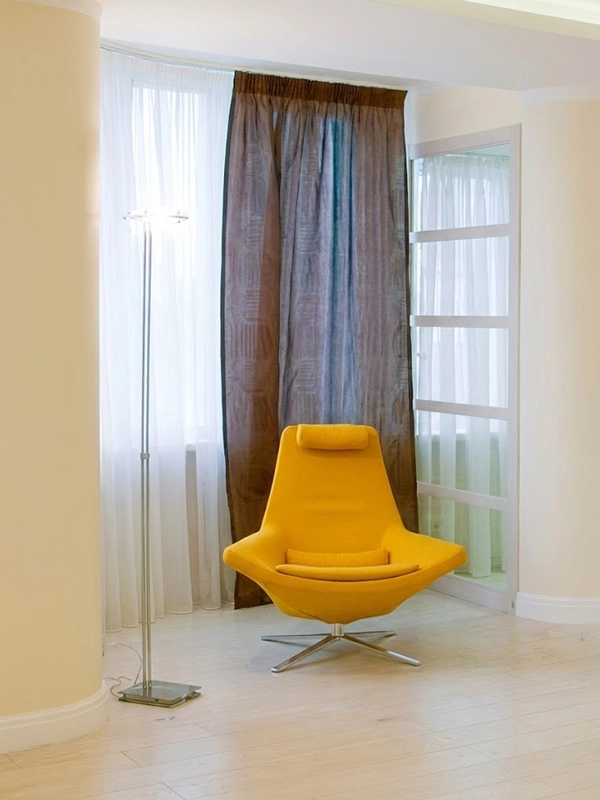 Интерьер светлой спальни с зеркальным панно в углу, плотными портьерами и мягким креслом с торшером